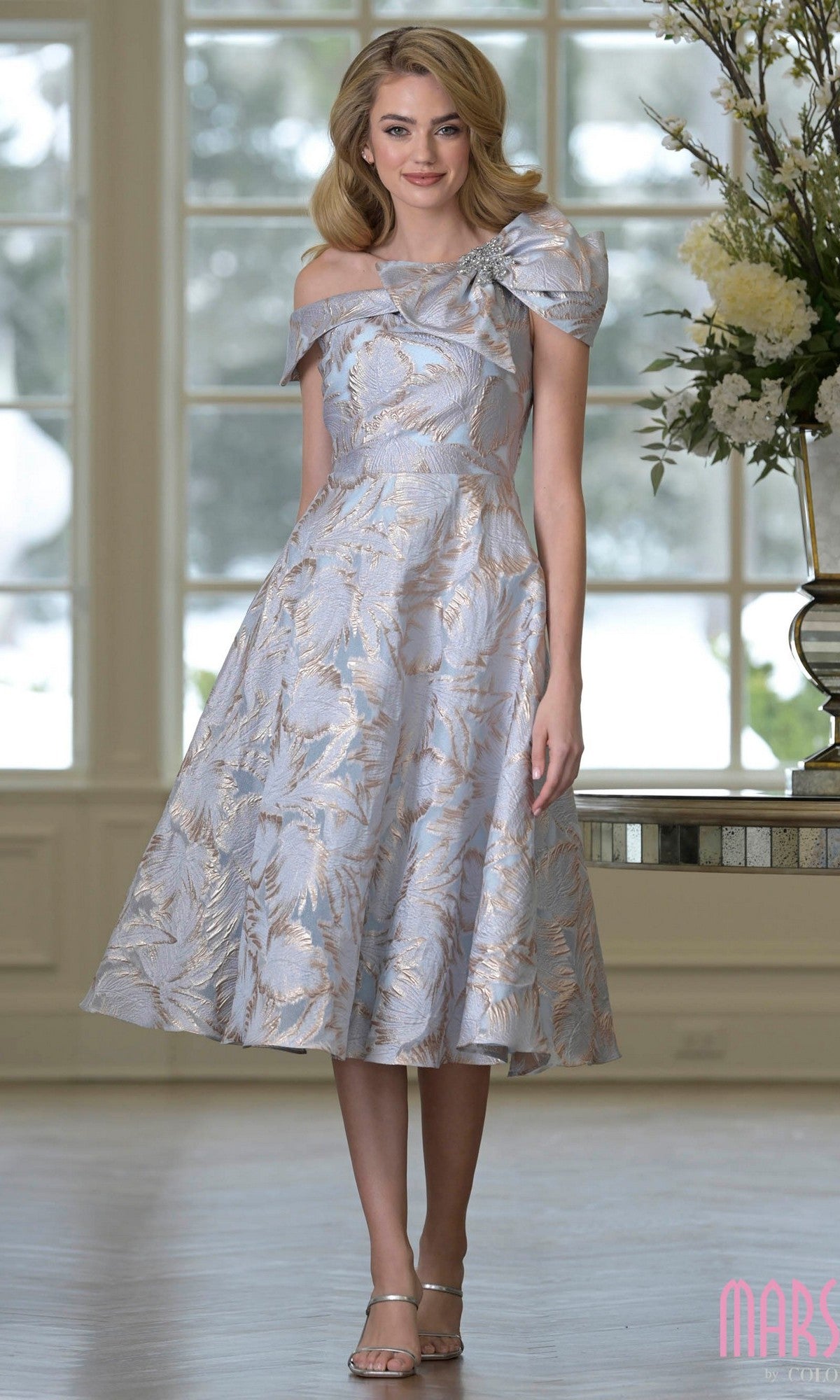 A woman stands indoors by a large window and floral arrangement, wearing the Marsoni MV1413 Knee Length Semi-Formal Dress in Gold Blue with off-the-shoulder style, textured jacquard fabric, and bow detail.